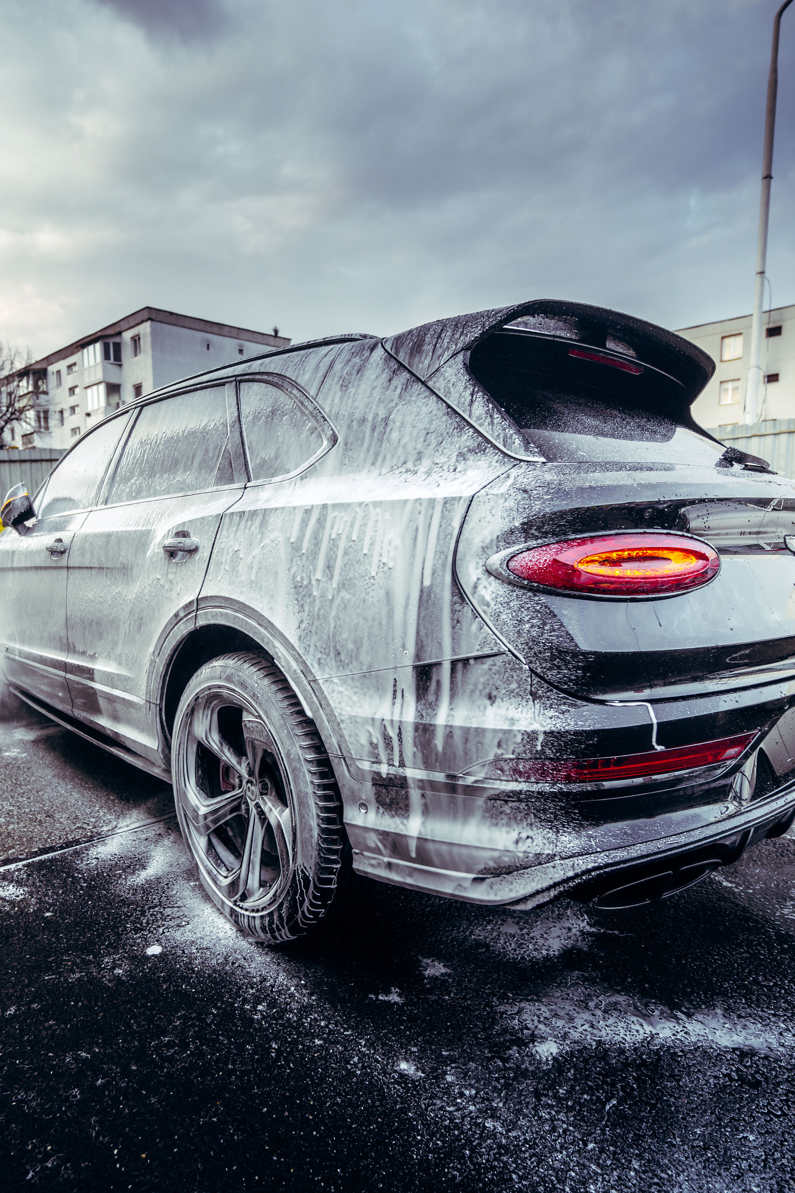 Grey Bentley Bentayga S covered in snow foam, seen from the rear three-quarter outdoors under an overcast sky