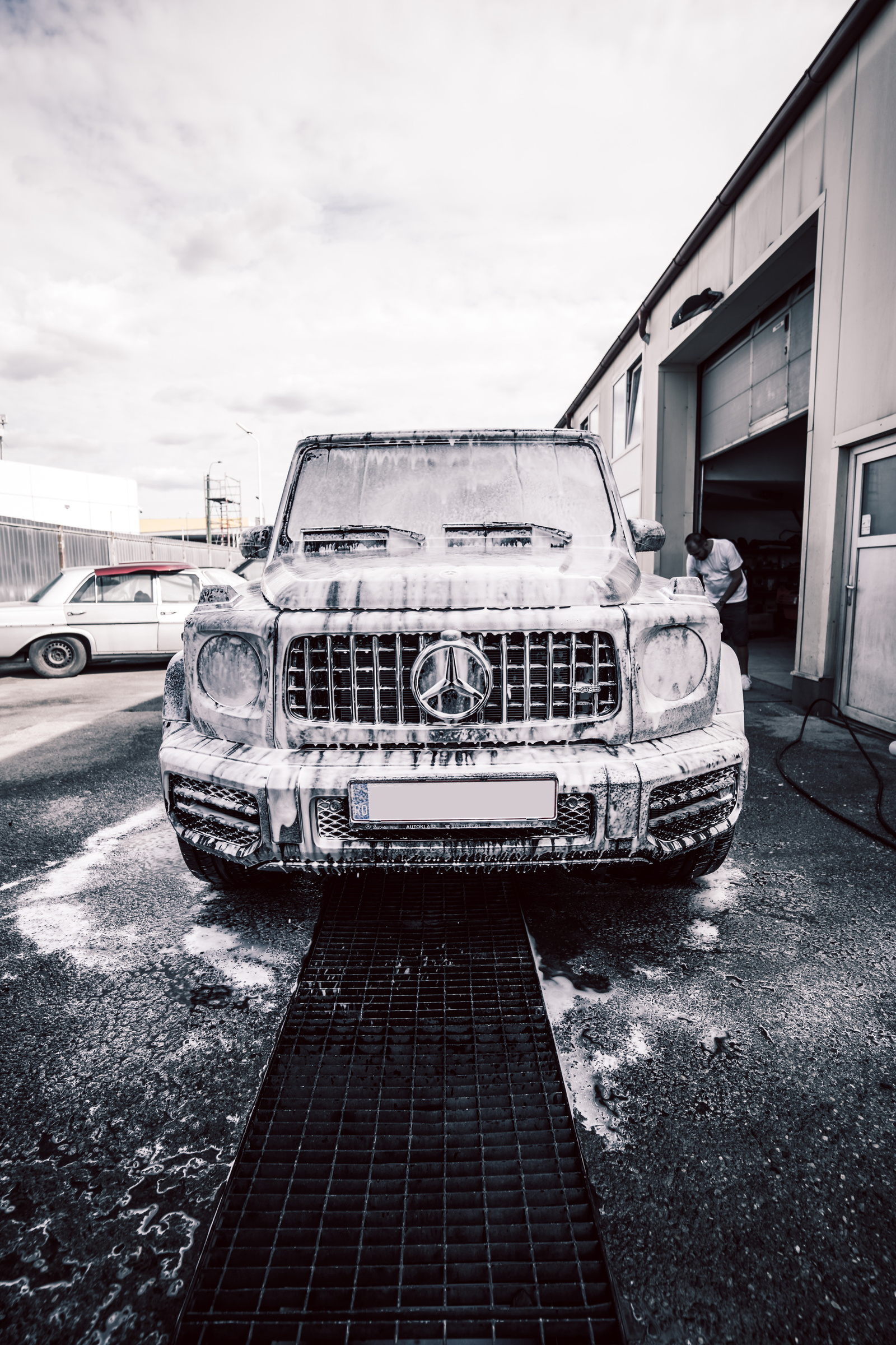 Mercedes-Benz G63 covered in snow foam, seen head-on over the wash grate outside a workshop, in a desaturated photo