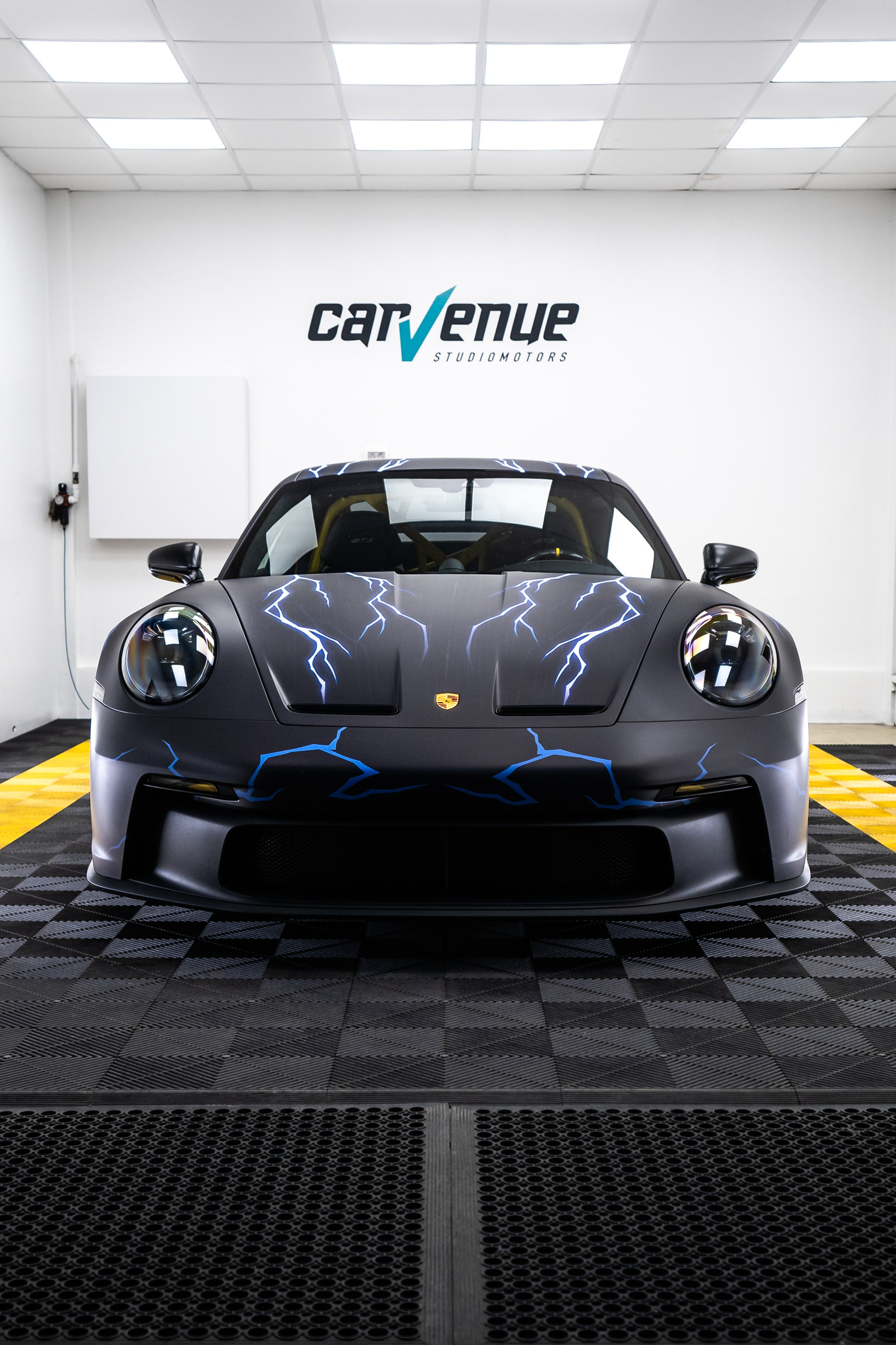 Front view of a Porsche 911 in a matte grey wrap with blue lightning bolts across the bonnet and front bumper, in the studio
