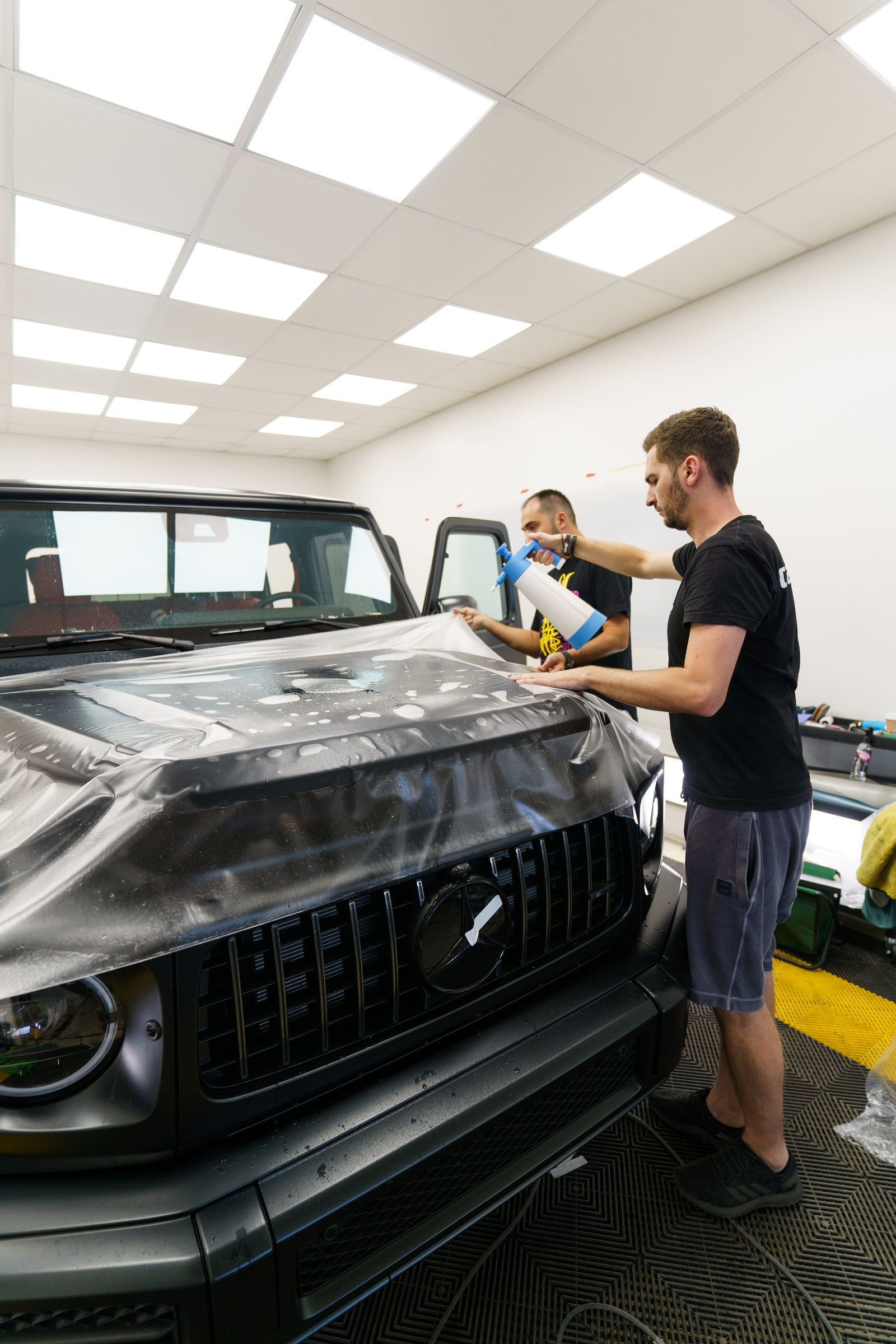 Two technicians lay clear paint protection film over the wet bonnet of a matte black Mercedes-AMG G-Class