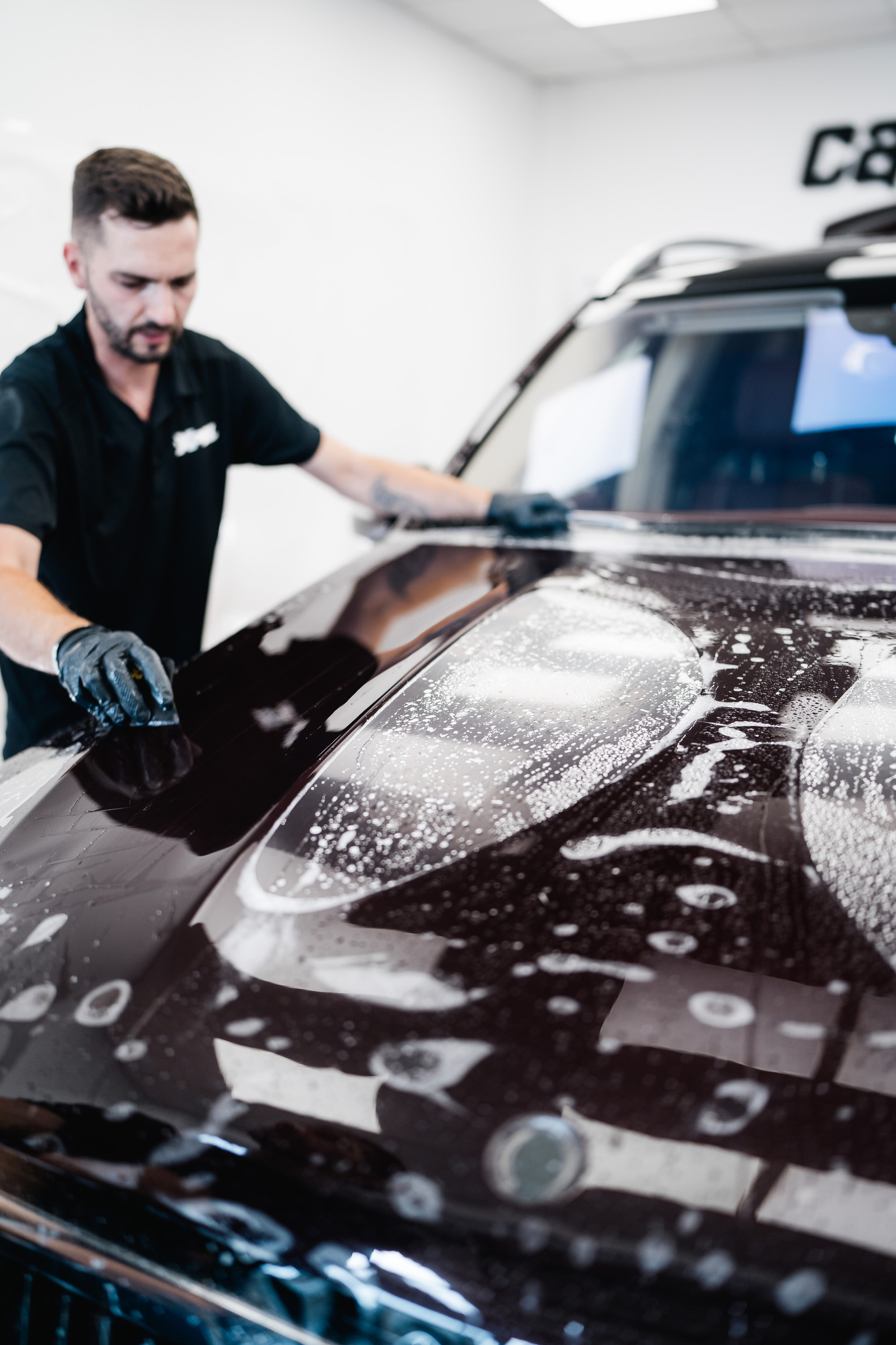 A technician smooths film with a squeegee across the wet bonnet of a dark SUV in the studio