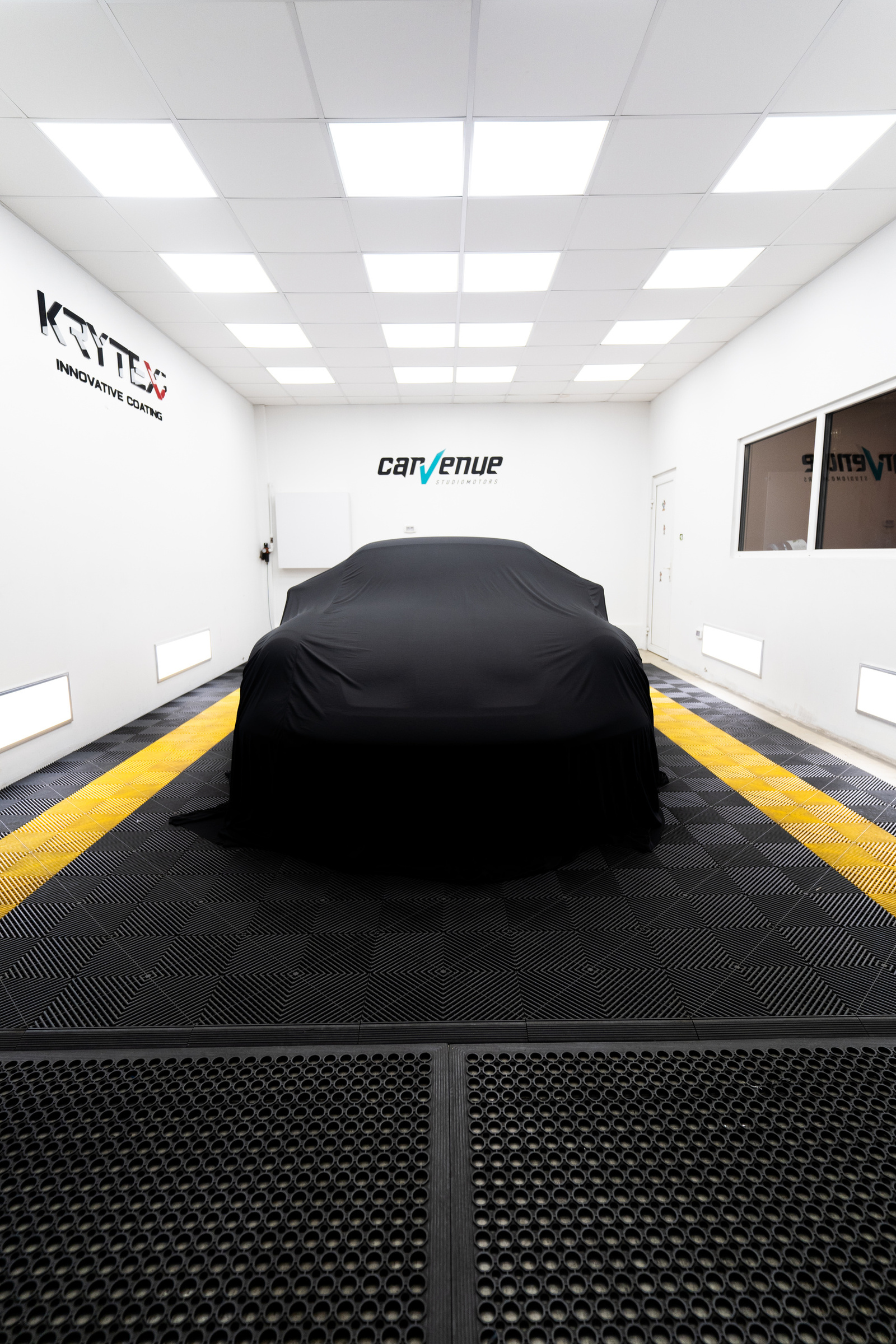 A car under a black cover in the CarVenue studio, with white walls and a black floor with yellow stripes