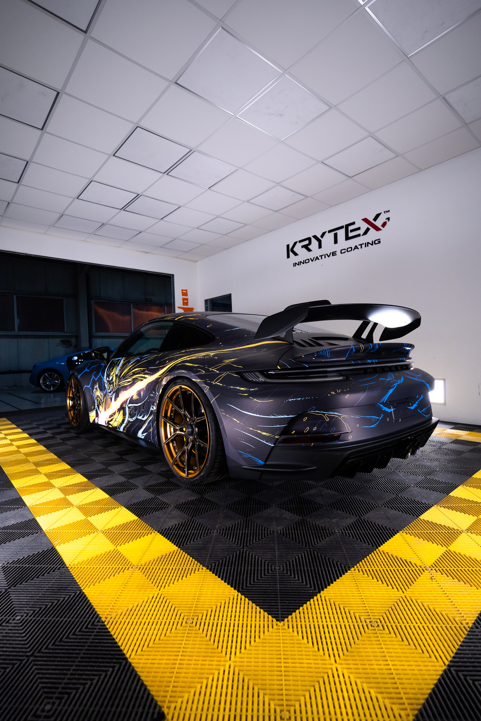 Rear view of a Porsche 911 in a matte wrap with yellow and blue comic-style artwork and gold wheels, in the studio