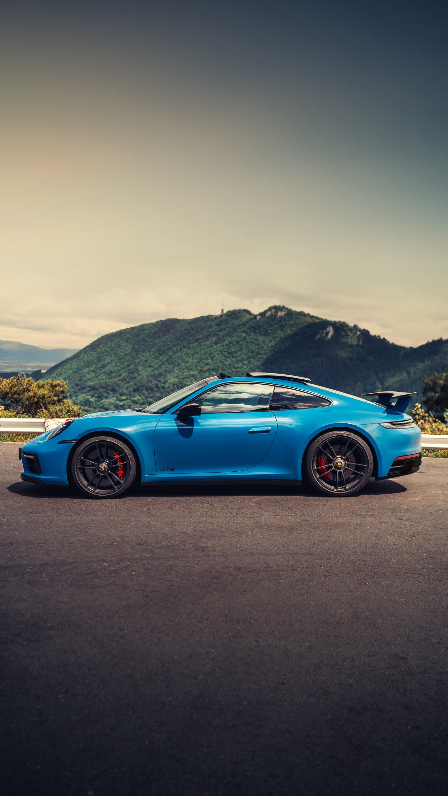 Porsche 911 GTS albastru, văzut din profil pe un drum de munte, cu dealuri împădurite în fundal