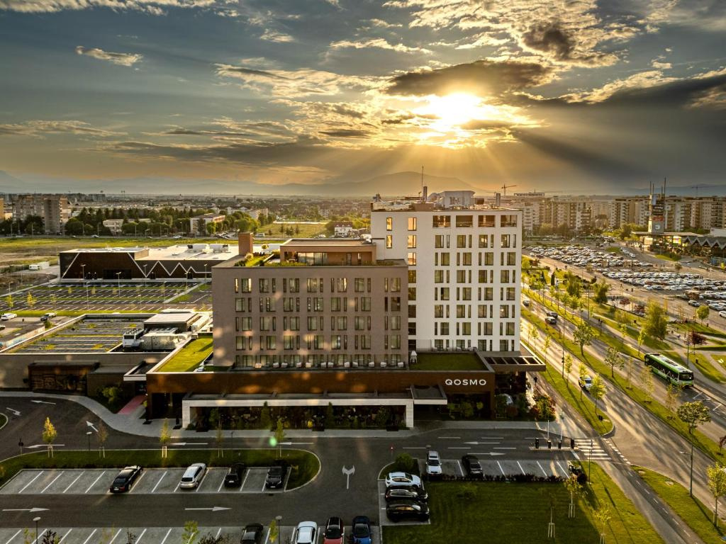 Hotel Qosmo Brașov from above, with the sun breaking through clouds, its car park in front and the city and mountains beyond