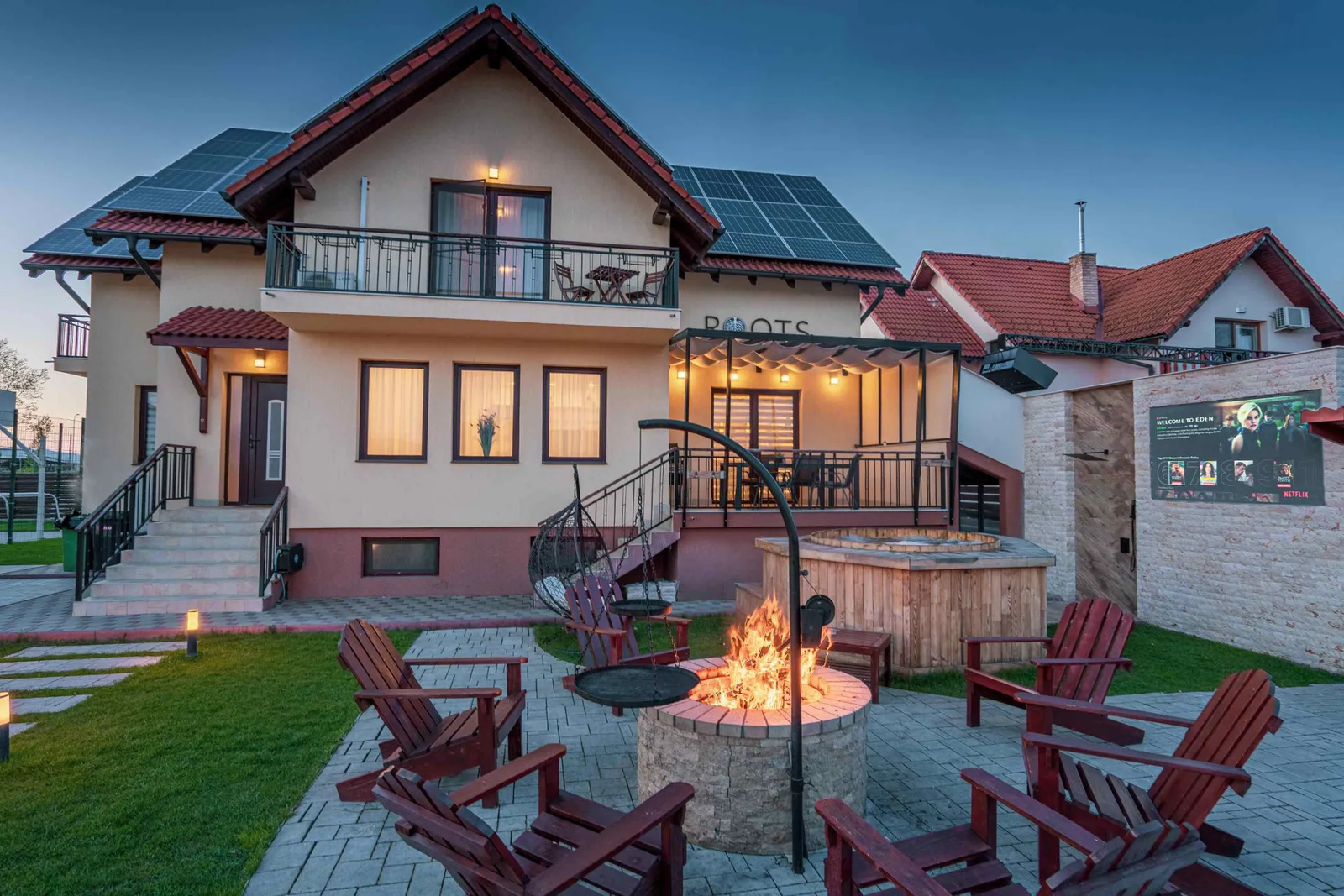 A ROOTS Villas house at dusk, with a fire burning in a stone fire pit, wooden chairs, a hot tub and an outdoor cinema screen