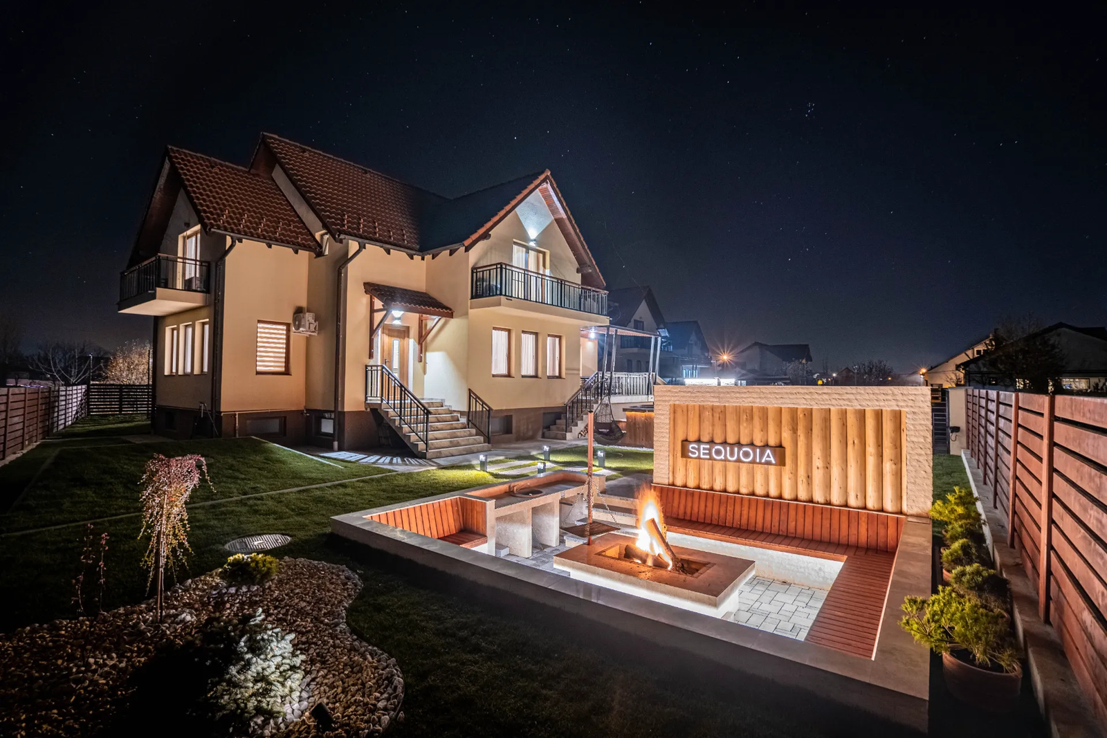 A ROOTS Villas house at night under a starry sky, with a sunken fire pit lounge and wooden benches in the garden